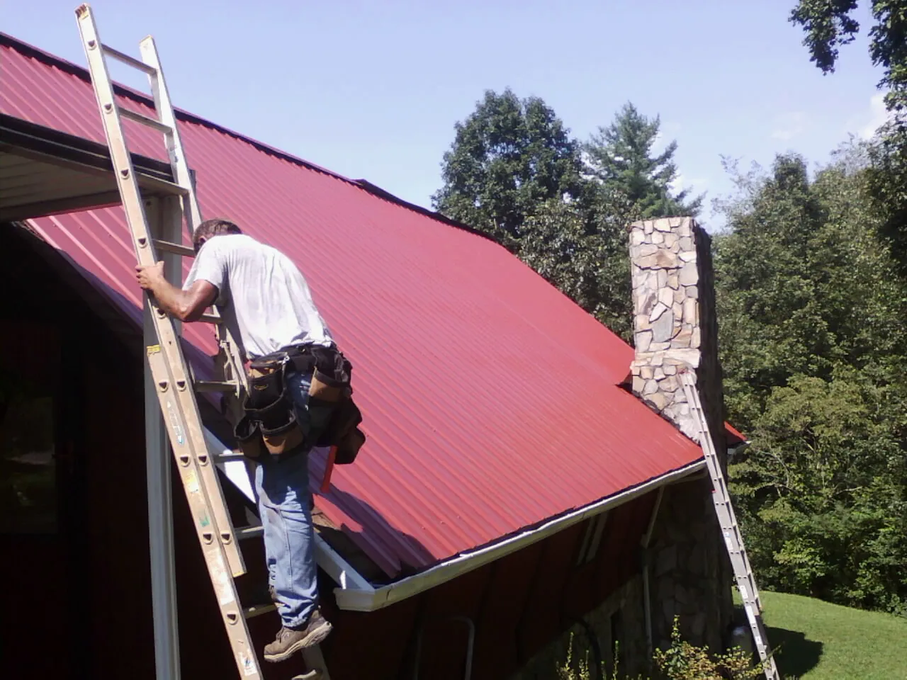 Roofer on a ladder beside a red metal roof and stone chimney — Soffit & Fascia Repair by Roofing Brothers in Casey